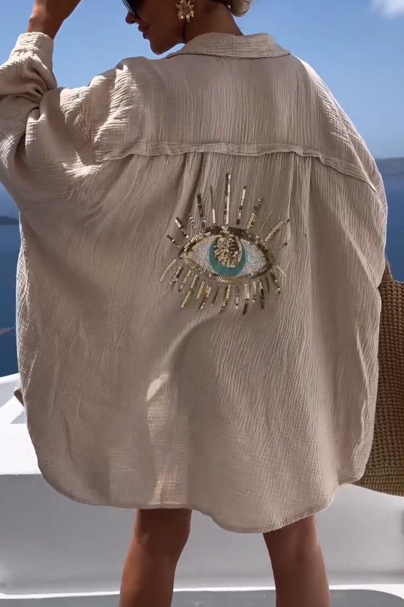 model draagt zomerse linnen blouse met lange mouwen katoenen shirt voor de zomer met lange mouwen, oversized strandtop met boze oog op de rug van glitter, vakantie outfit beige